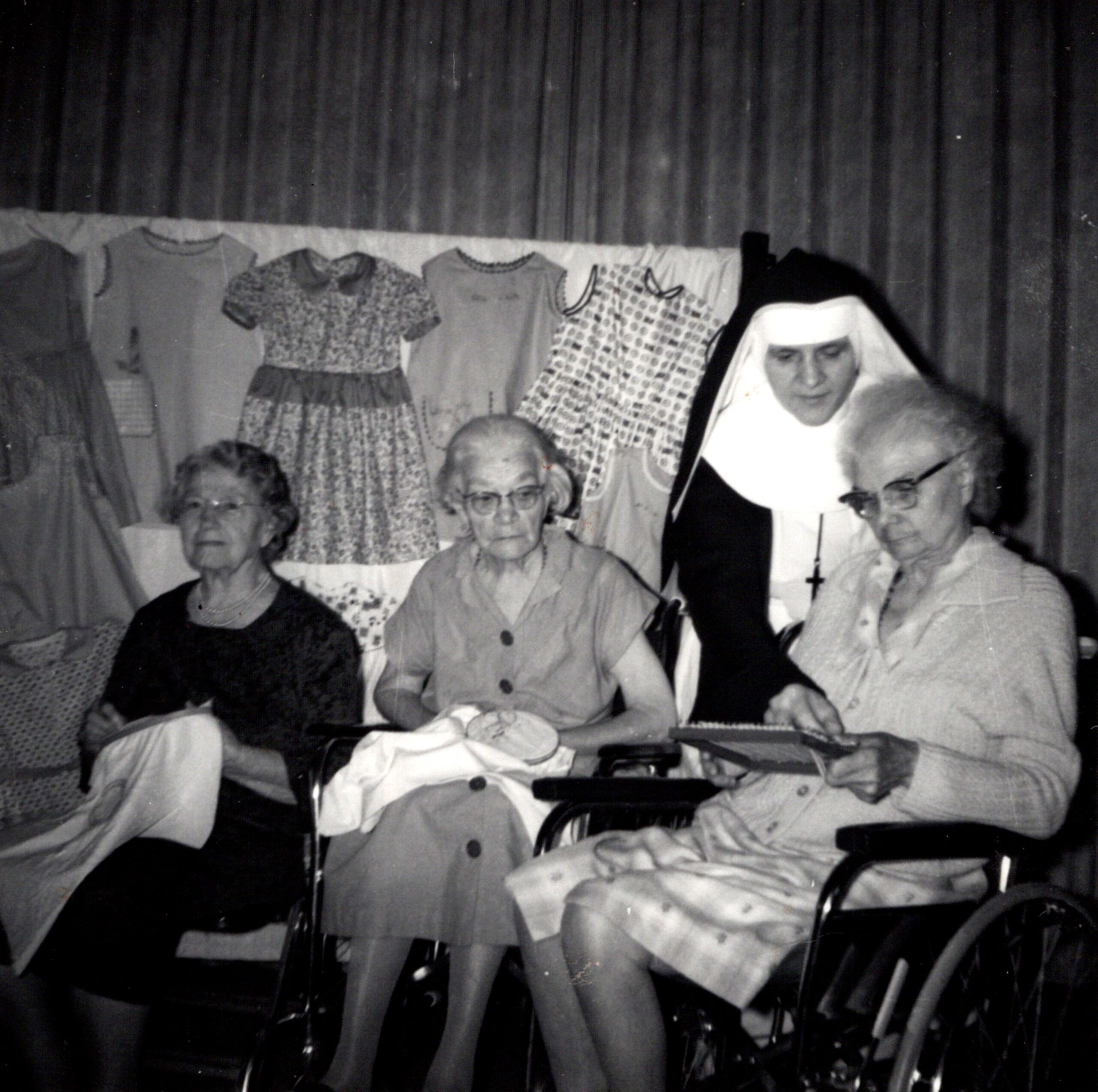 An old black and white photograph of residents at the Home many years ago. Three female residents are cross-stitching or sewing, and a Sister in a habit is helping them.