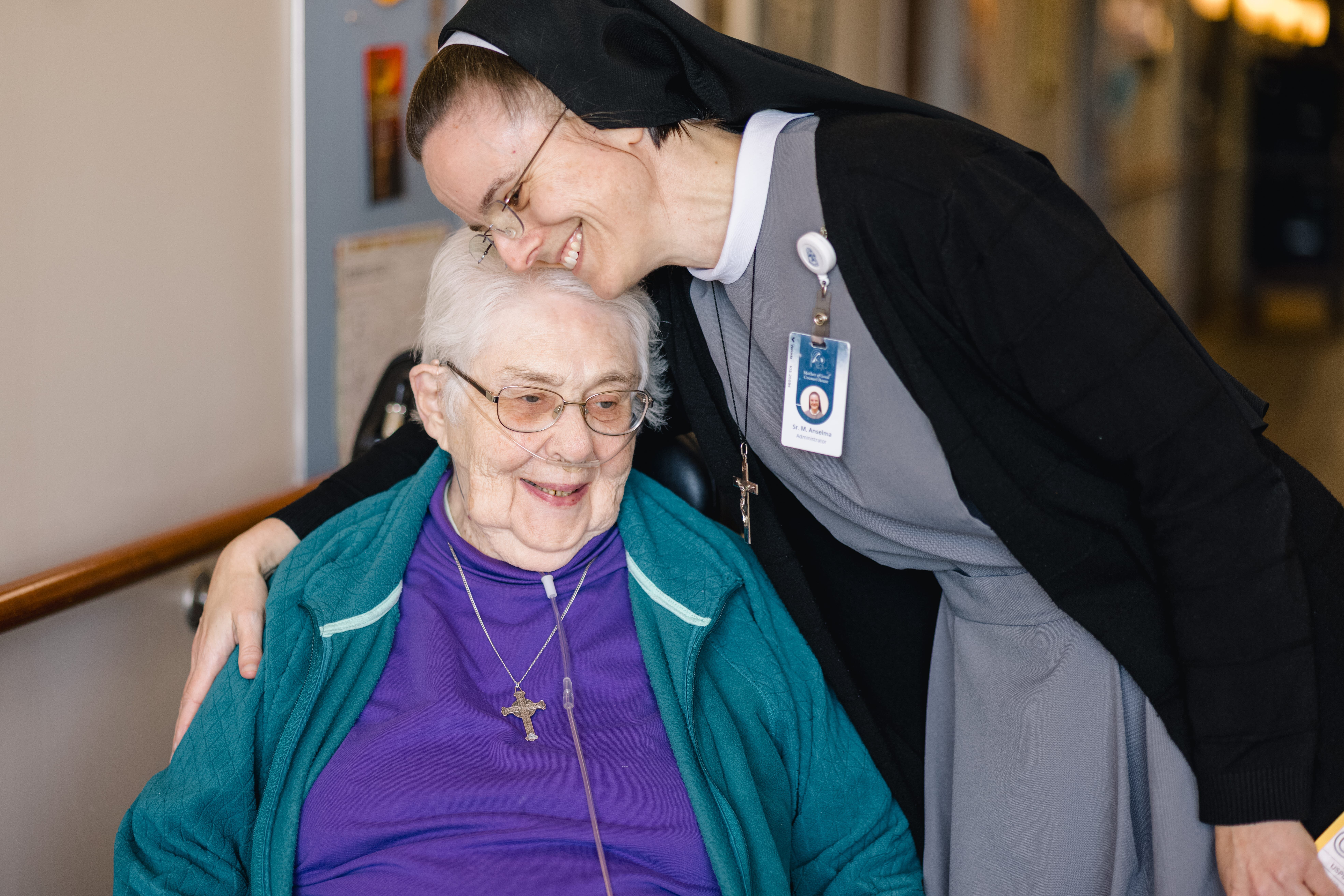 MOGC10.21.25-149 Sister M. Anselma hugging a resident in a wheelchair with a cross around both of their necks. The resident has a cannula in her nose.