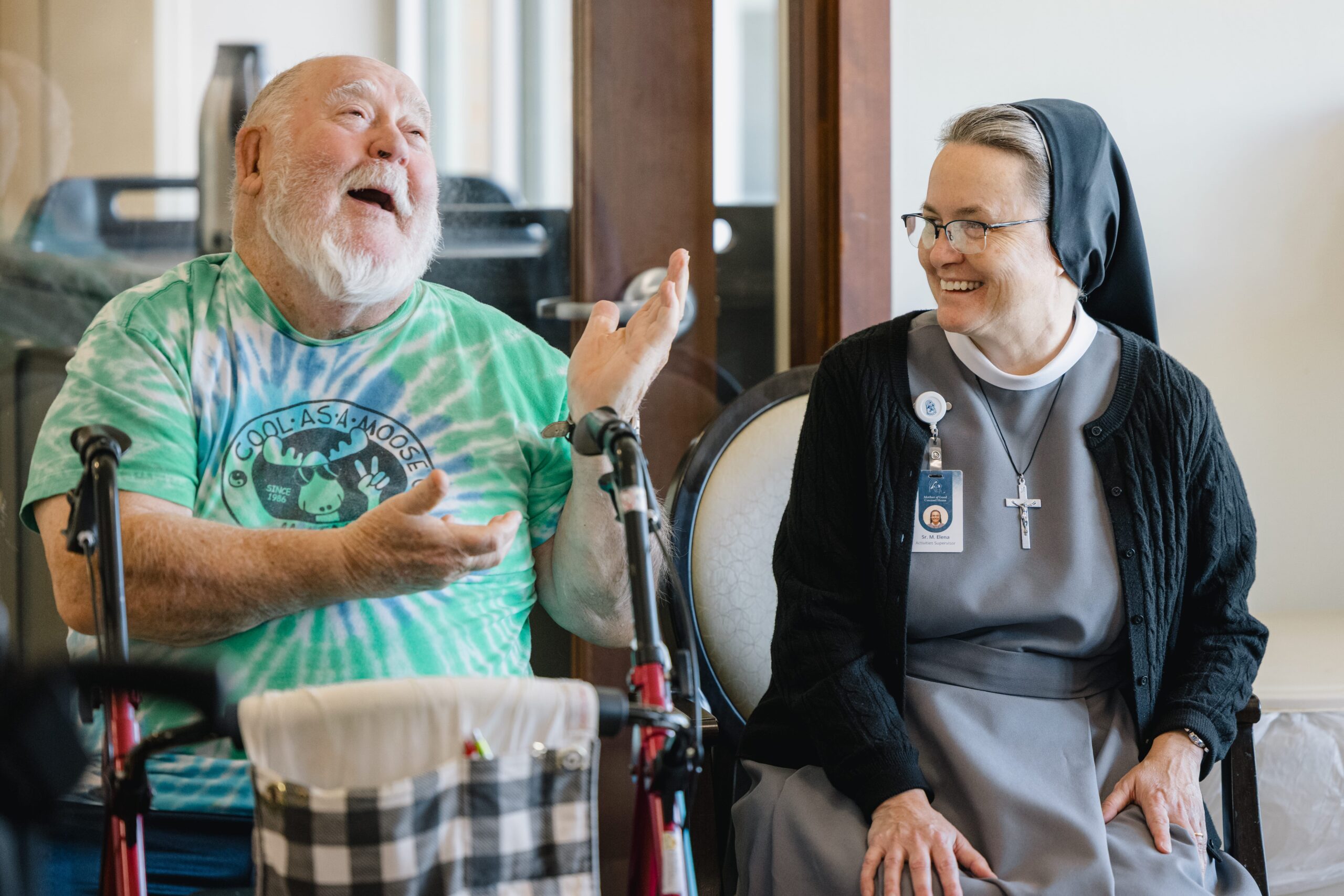 MOGC10.21.25-41 A resident and a Sister sitting together, joking around. The resident has a walker.