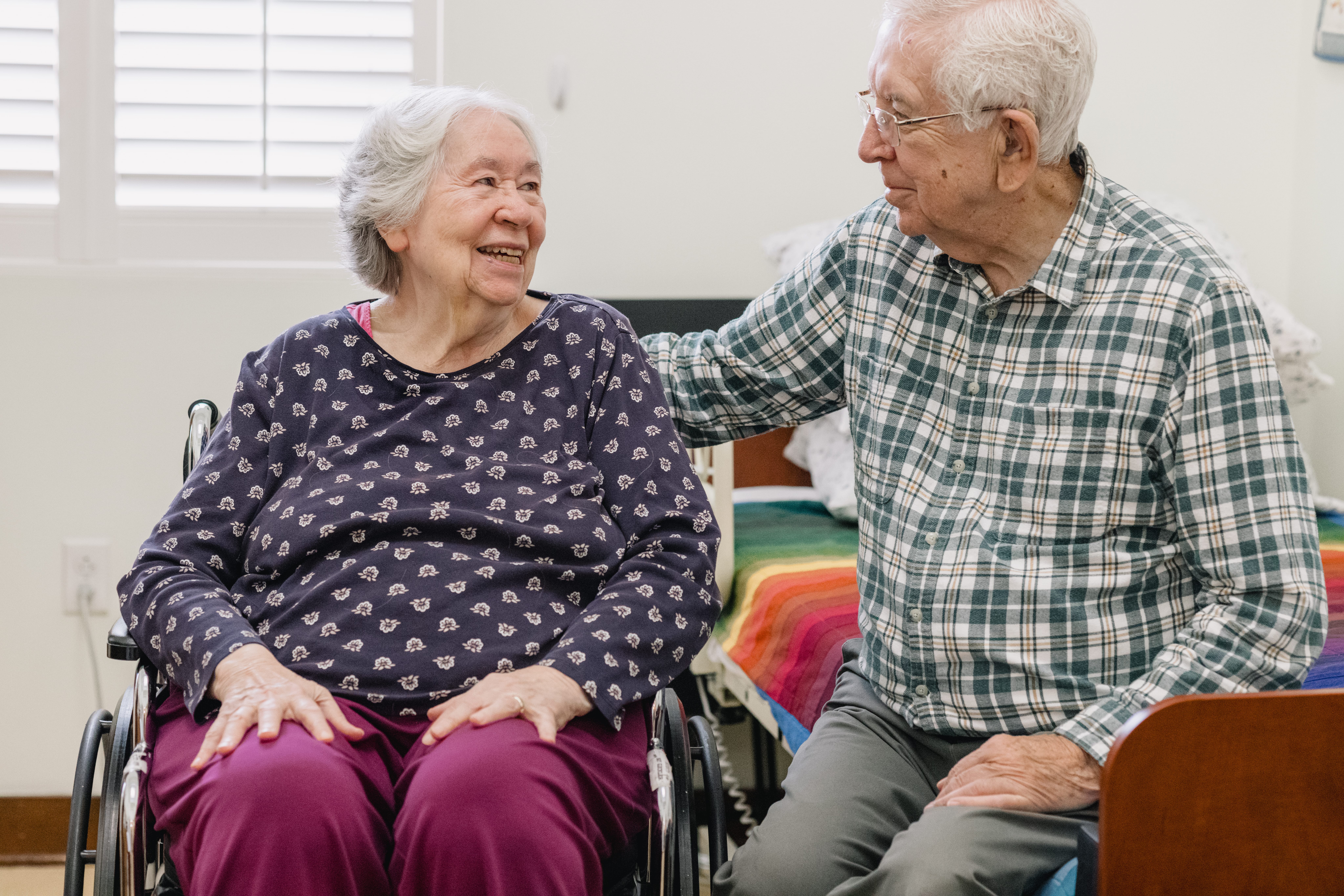 A resident and her spouse sitting together in her room. She is in her wheelchair, and he is sitting on the bed.