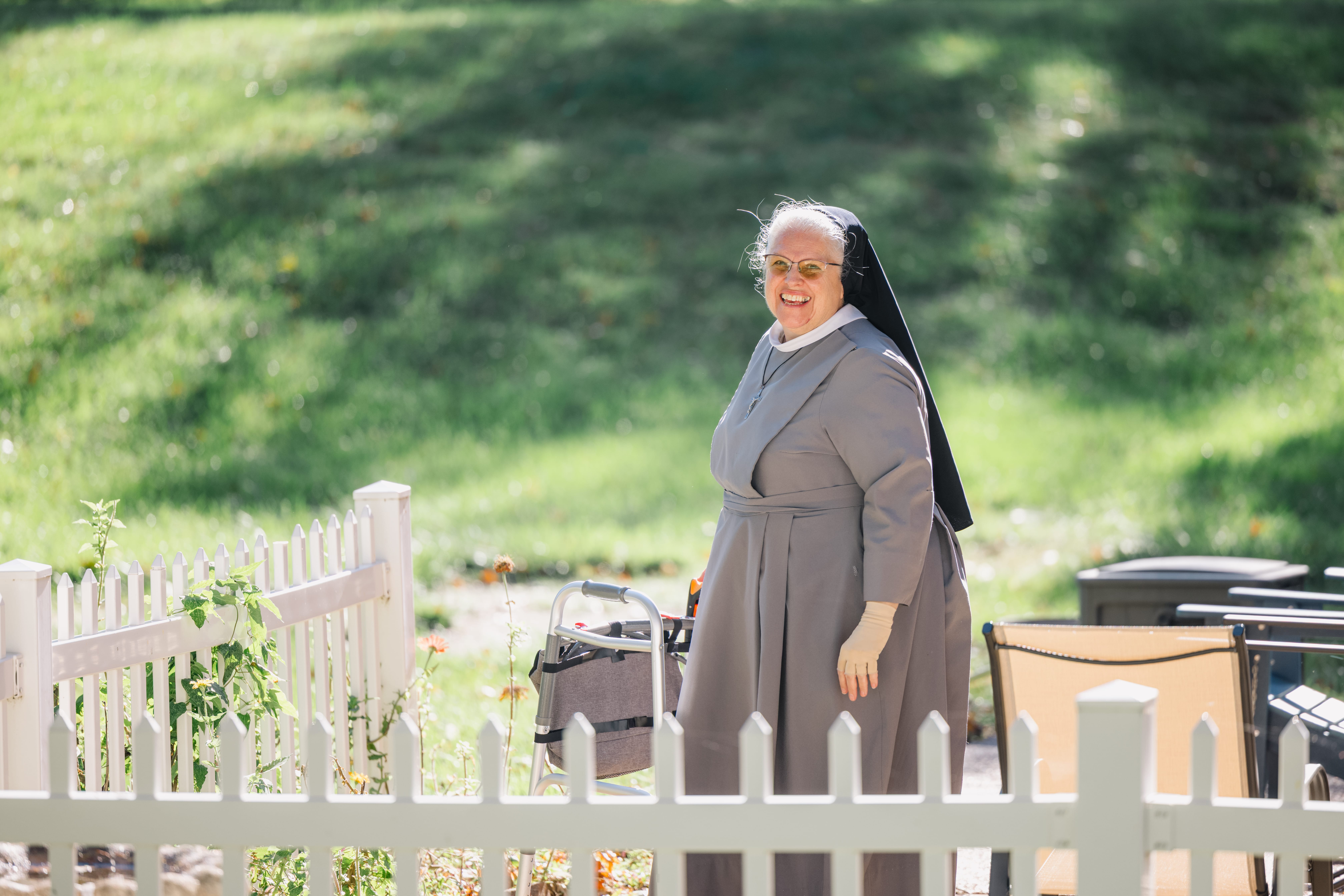 Sister M. Jessica stands out in the backyard behind a picket fence on a sunny day. She's smiling and standing with her walker.