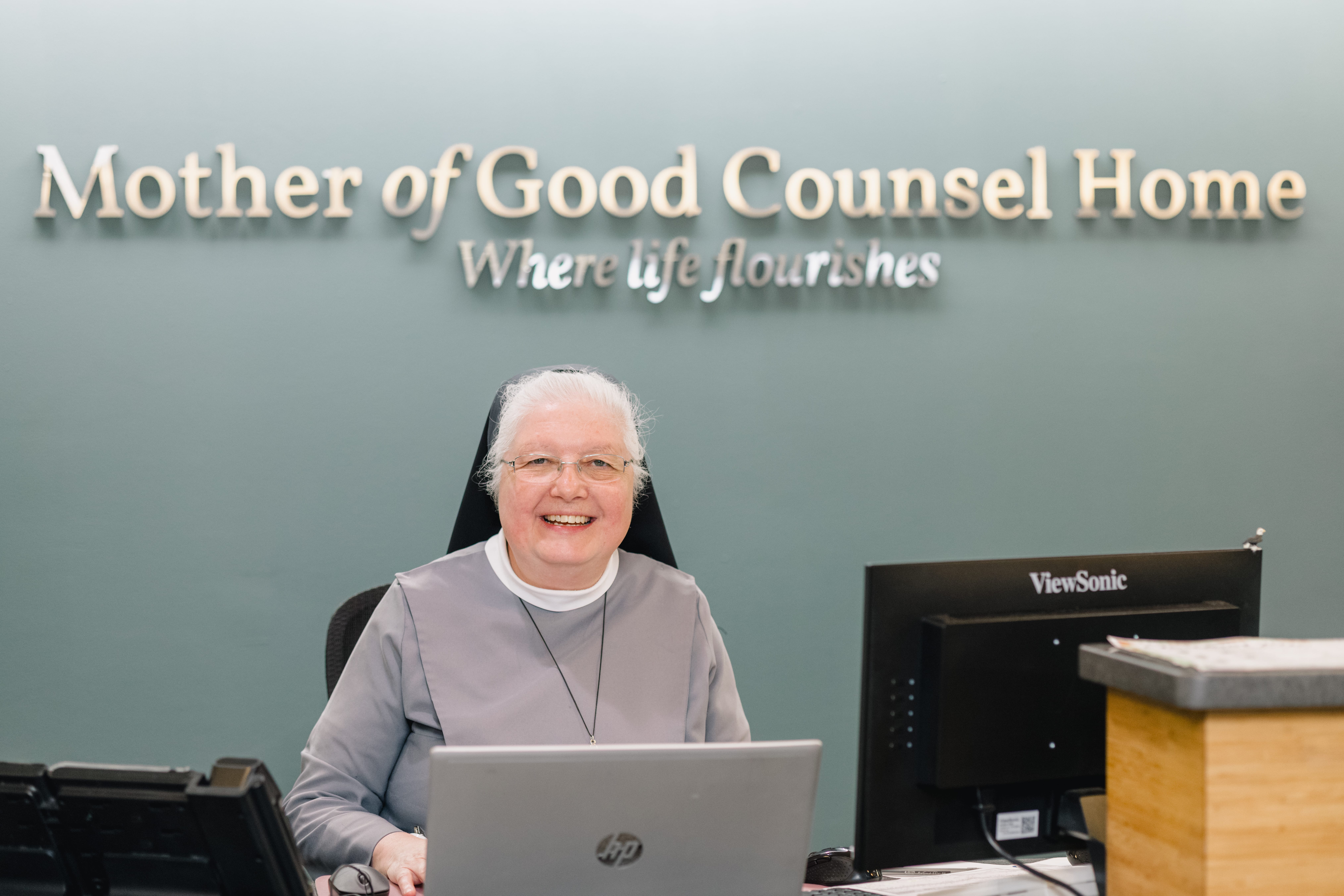 Sister M. Clara sitting at the front desk behind a computer, smiling. There is a Mother of Good Counsel Home sign behind her that also reads "Where Life Flourishes" below it.