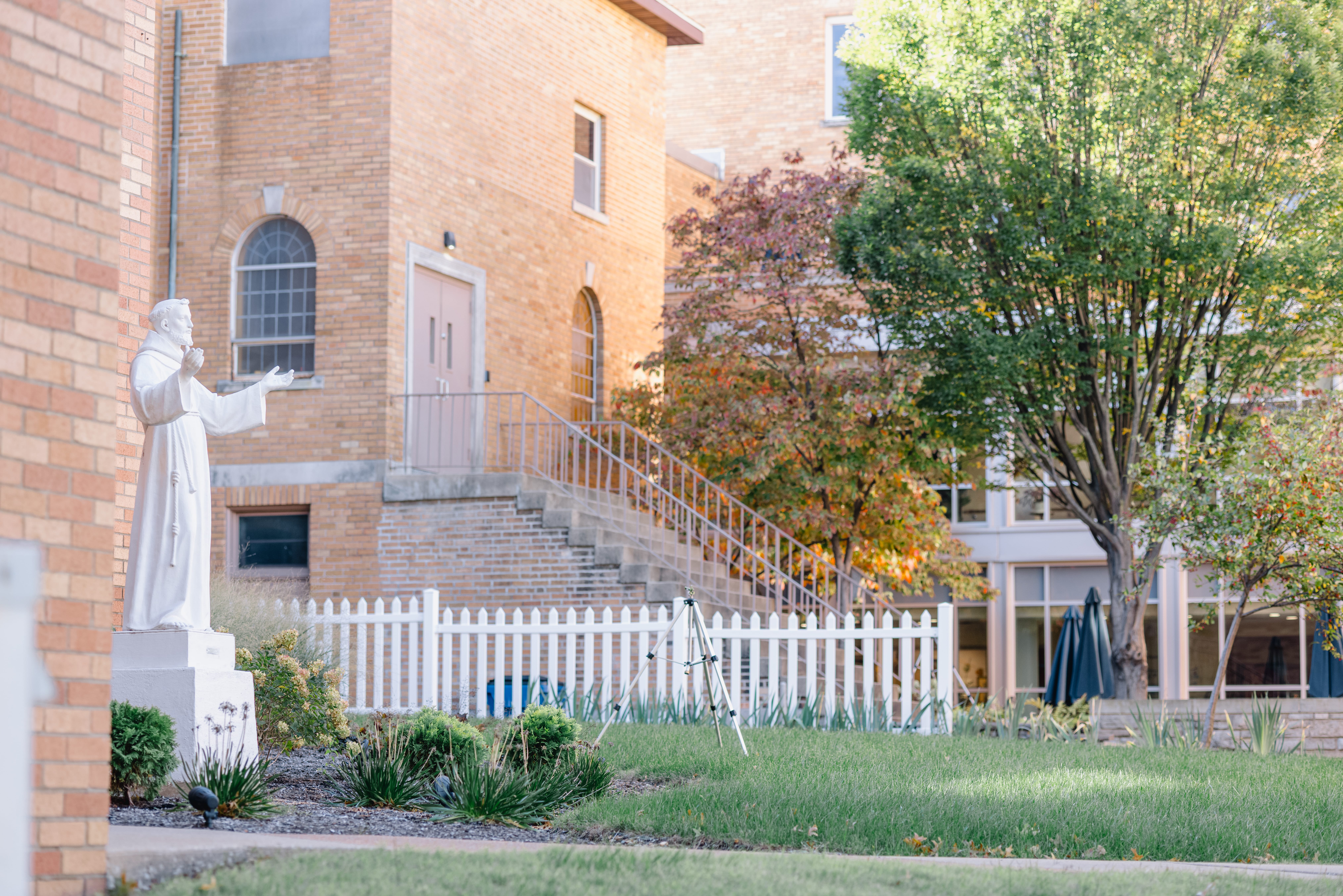 The Home's backyard. There is a statue of St. Francis, trees changing to fall colors, and steps up to the back of the chapel.