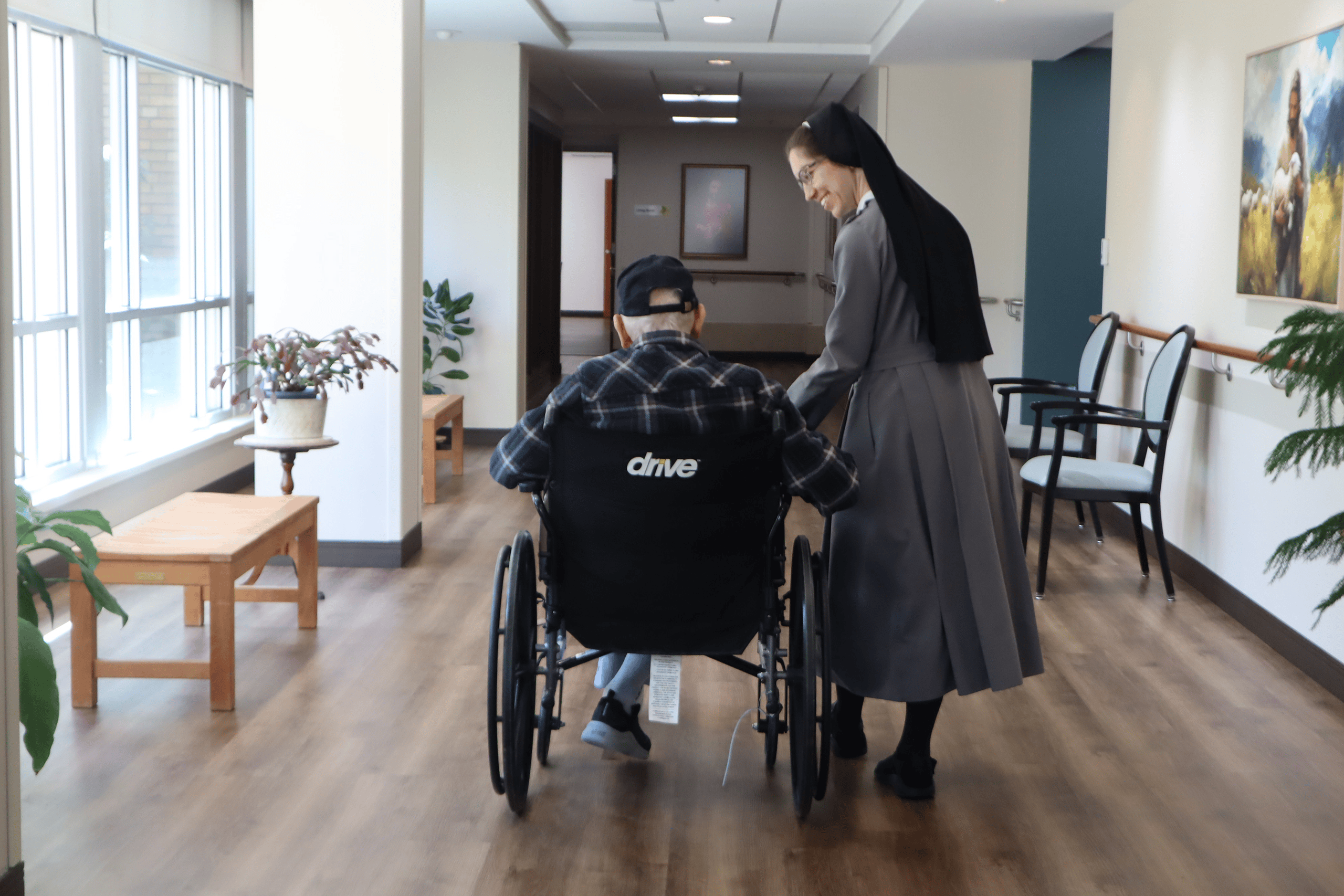 Sister M. Caterina walking with a resident in a wheelchair through a bright hallway.