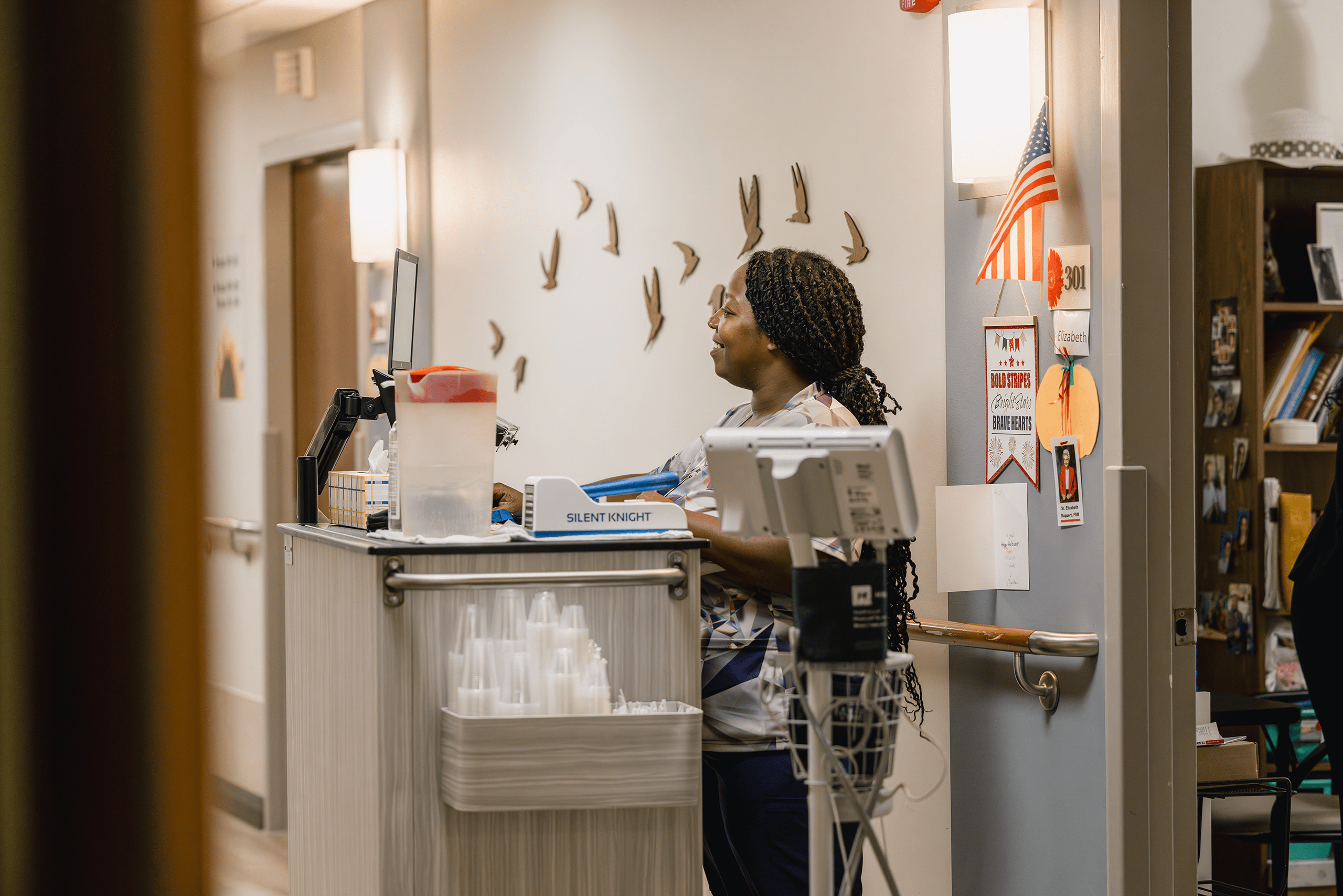 A Mission Partner standing outside a resident's room looking at her computer and preparing to administer medication.