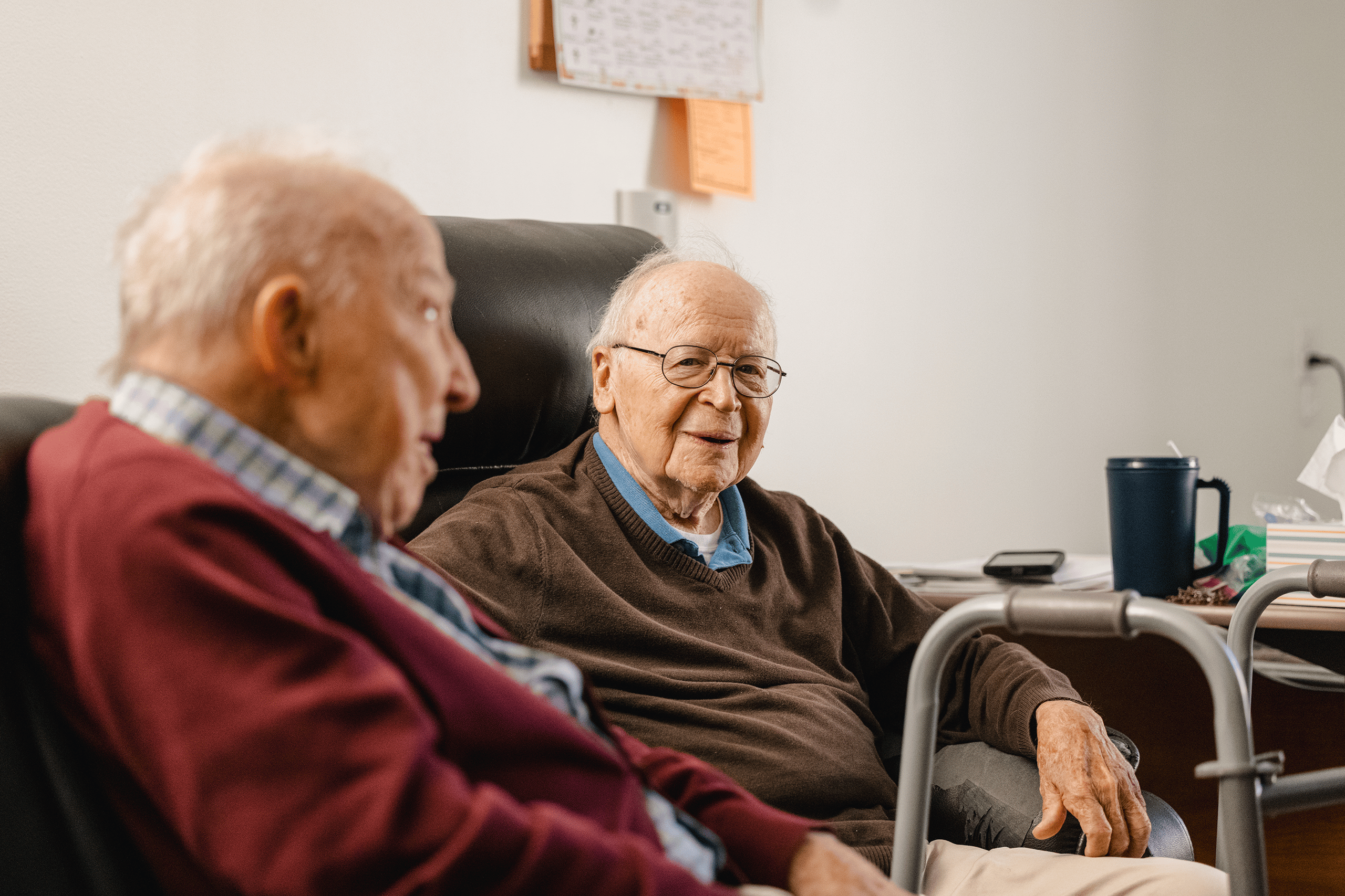 Two residents visiting together in a resident room. One is in his recliner. There is a walker in front of him, and a cup of water next to him.