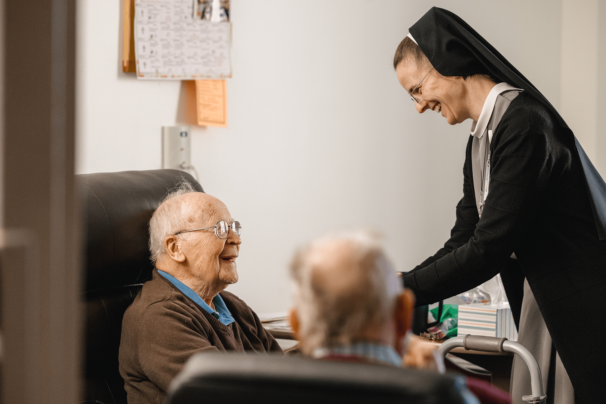 Sister M. Anselma greeting a resident in his room. He is sitting in his recliner smiling, and another resident is visiting with them in a different chair.