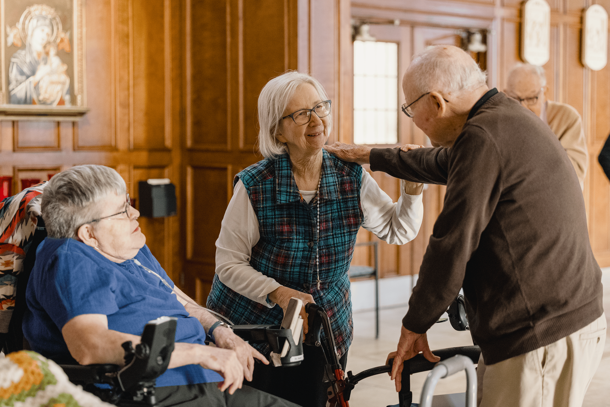 A resident greeting two other residents after mass. One is in a wheelchair, and two are on walkers. The resident has his hand on her shoulder and everyone is smiling.