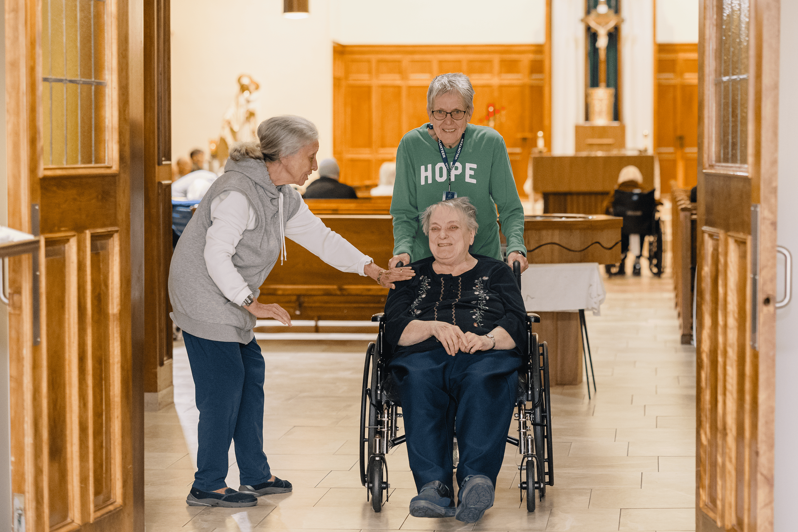 A resident is being pushed by a volunteer out of the Chapel after mass, and is being greeted by another resident as they pass by.